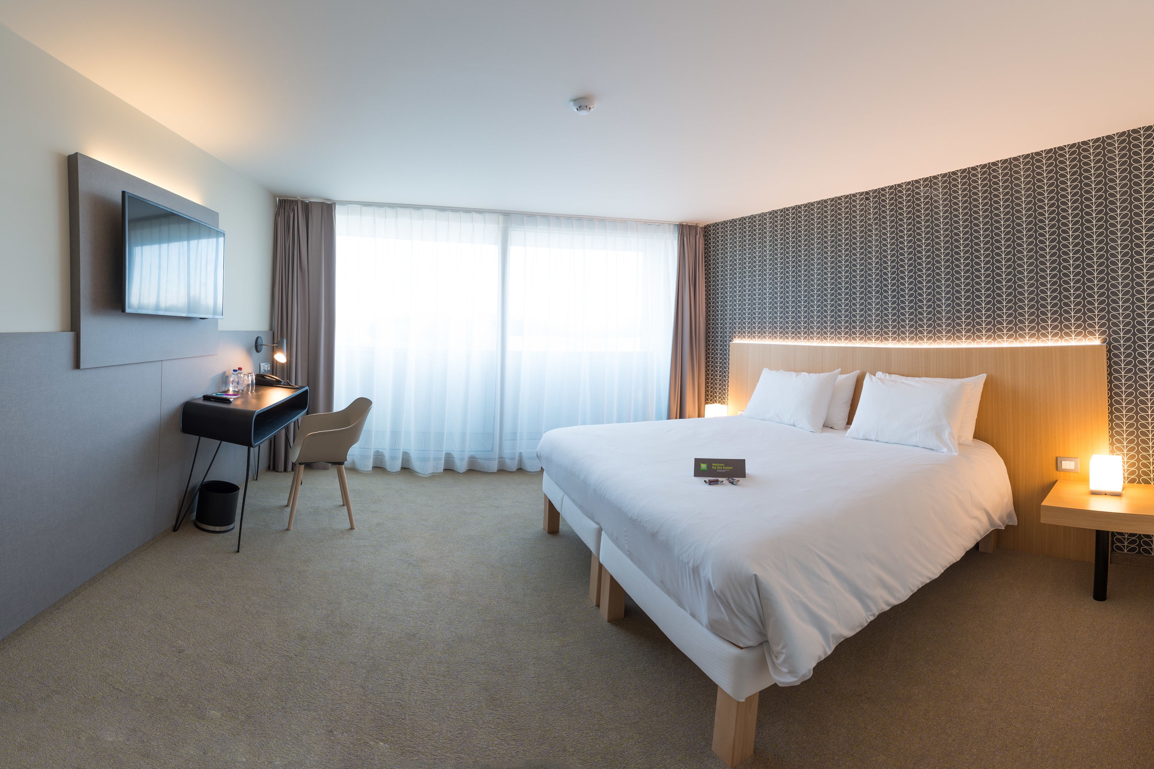 A hotel room with a large bed, a desk with a chair, a flat-screen TV, and a large window with sheer curtains.