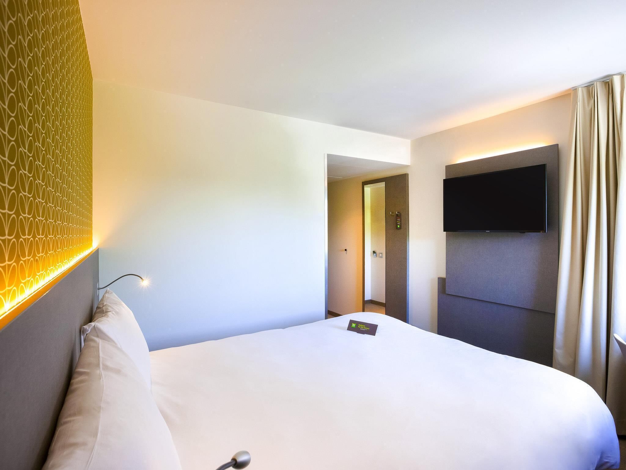 Habitación de hotel con cama, televisor montado en la pared y puerta que da a un pasillo.