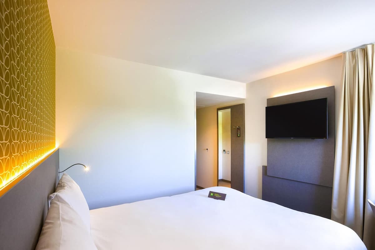 Quarto de hotel com uma cama, uma televisão fixada na parede e uma porta que dá para um corredor.