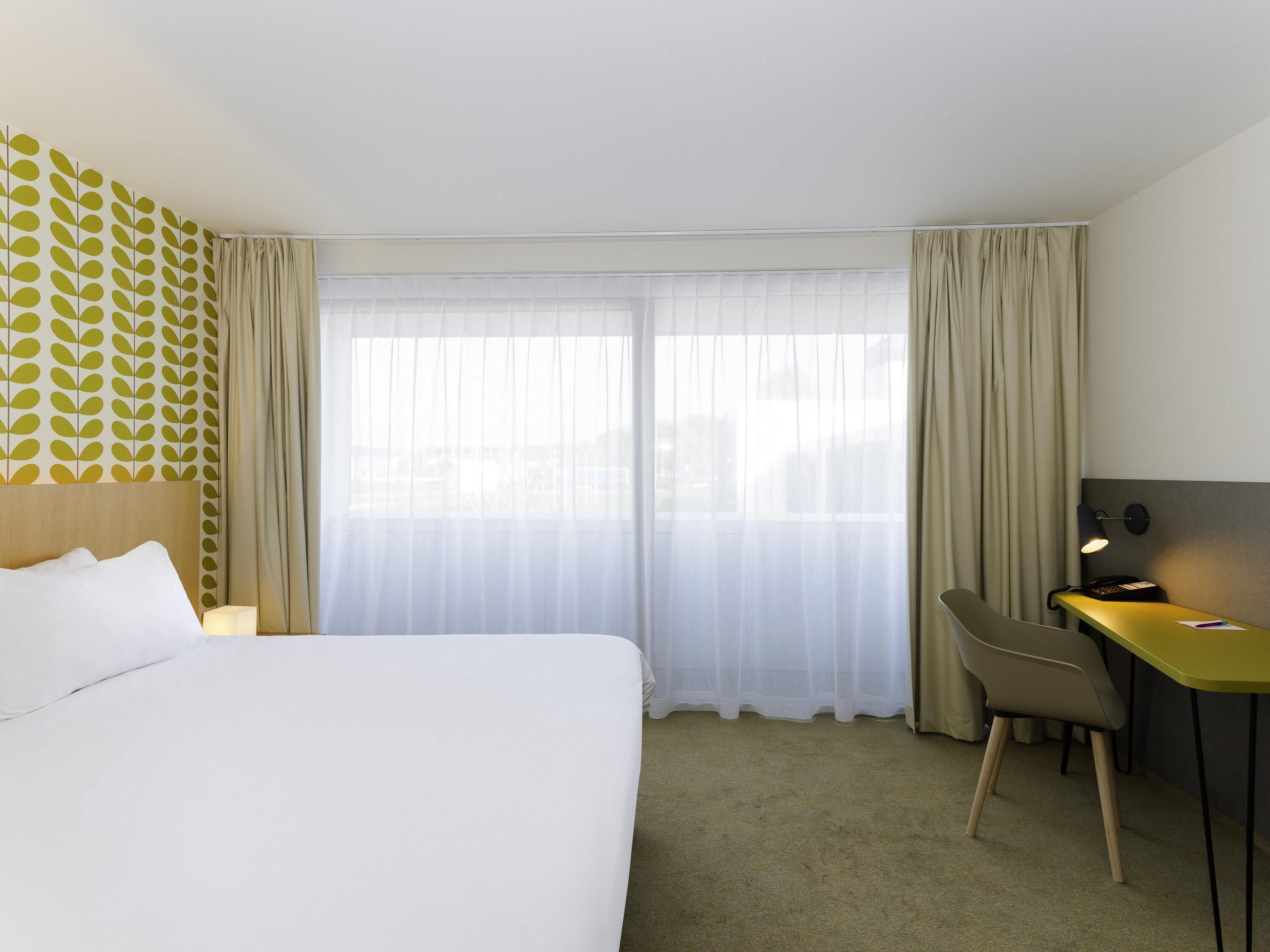 Quarto de hotel com cama, escrivaninha, cadeira, luminária e janela com cortinas translúcidas.