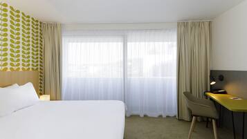 Quarto de hotel com cama, escrivaninha, cadeira, luminária e janela com cortinas translúcidas.