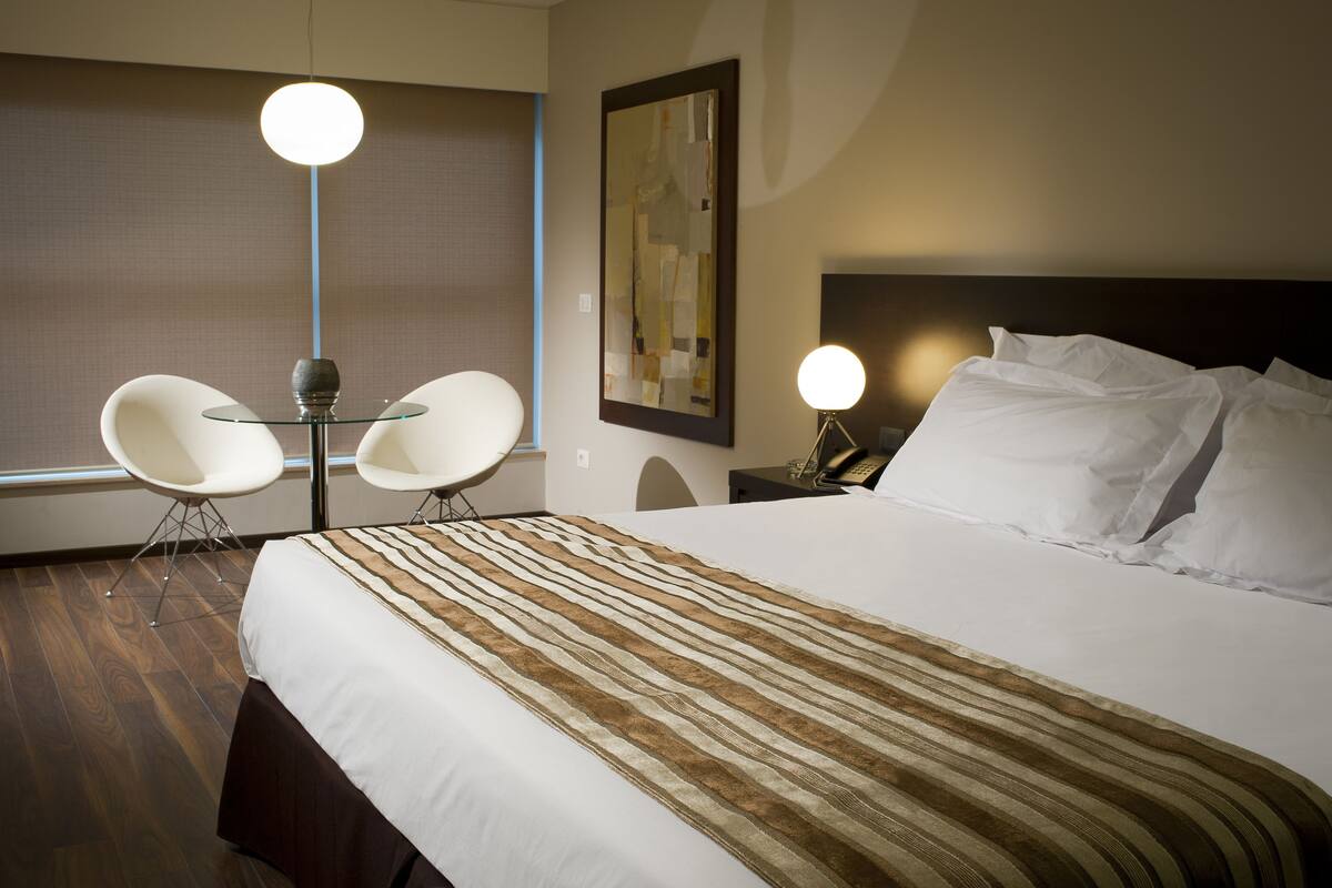 Une chambre d’hôtel avec un grand lit, deux chaises blanches, une table en verre et une lampe moderne.