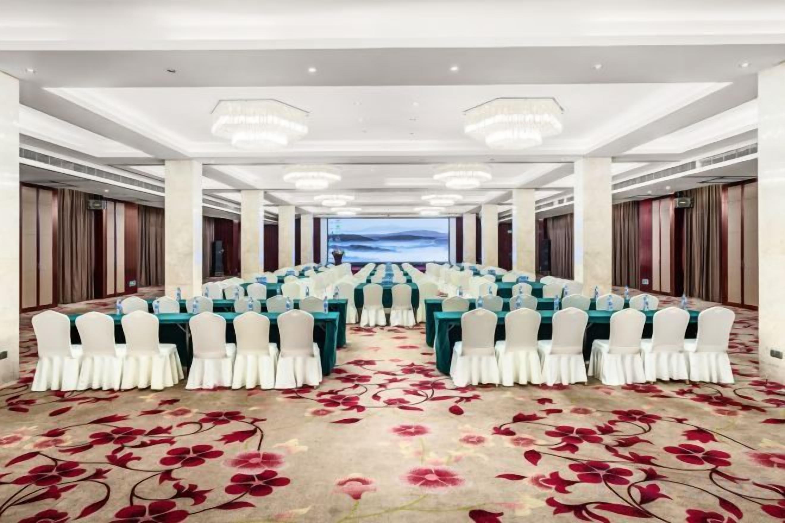 A conference room with rows of tables and chairs, a carpet with a floral pattern, and a view of the sky through a window.