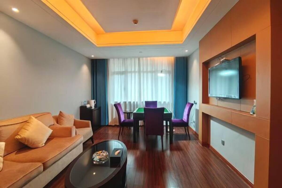 A hotel room with a sofa, a coffee table, a TV mounted on the wall, a dining table with chairs, and a large window with curtains.