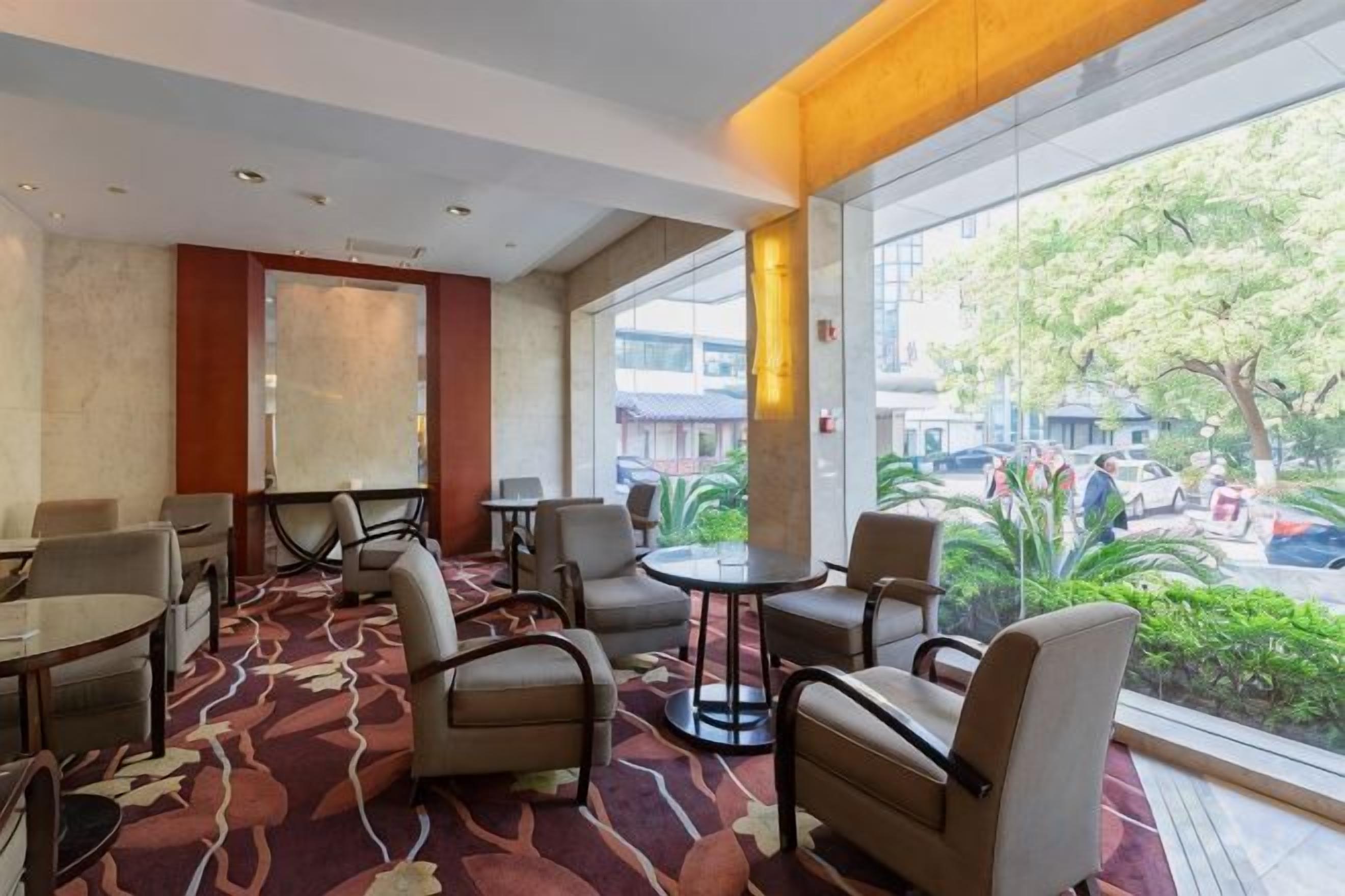 A hotel lobby with seating arrangements, a large window offering a view of the outdoors, and a patterned carpet.