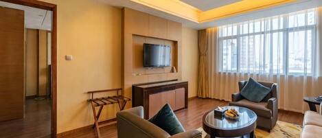 A hotel room with a flat-screen TV mounted on the wall, a sofa, a coffee table, and a large window providing natural light.