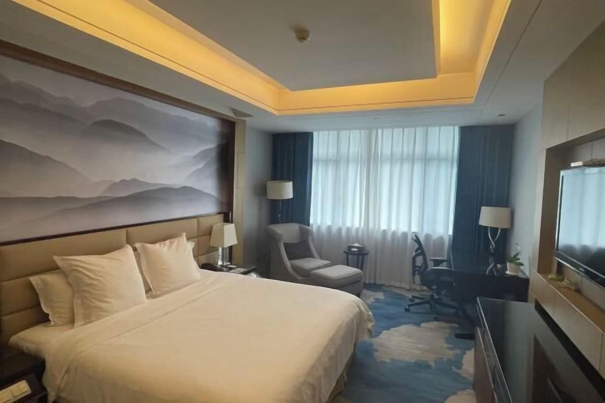 A hotel room with a large bed, two bedside tables with lamps, a desk with a chair, a large flat-screen TV, and a ceiling mural.
