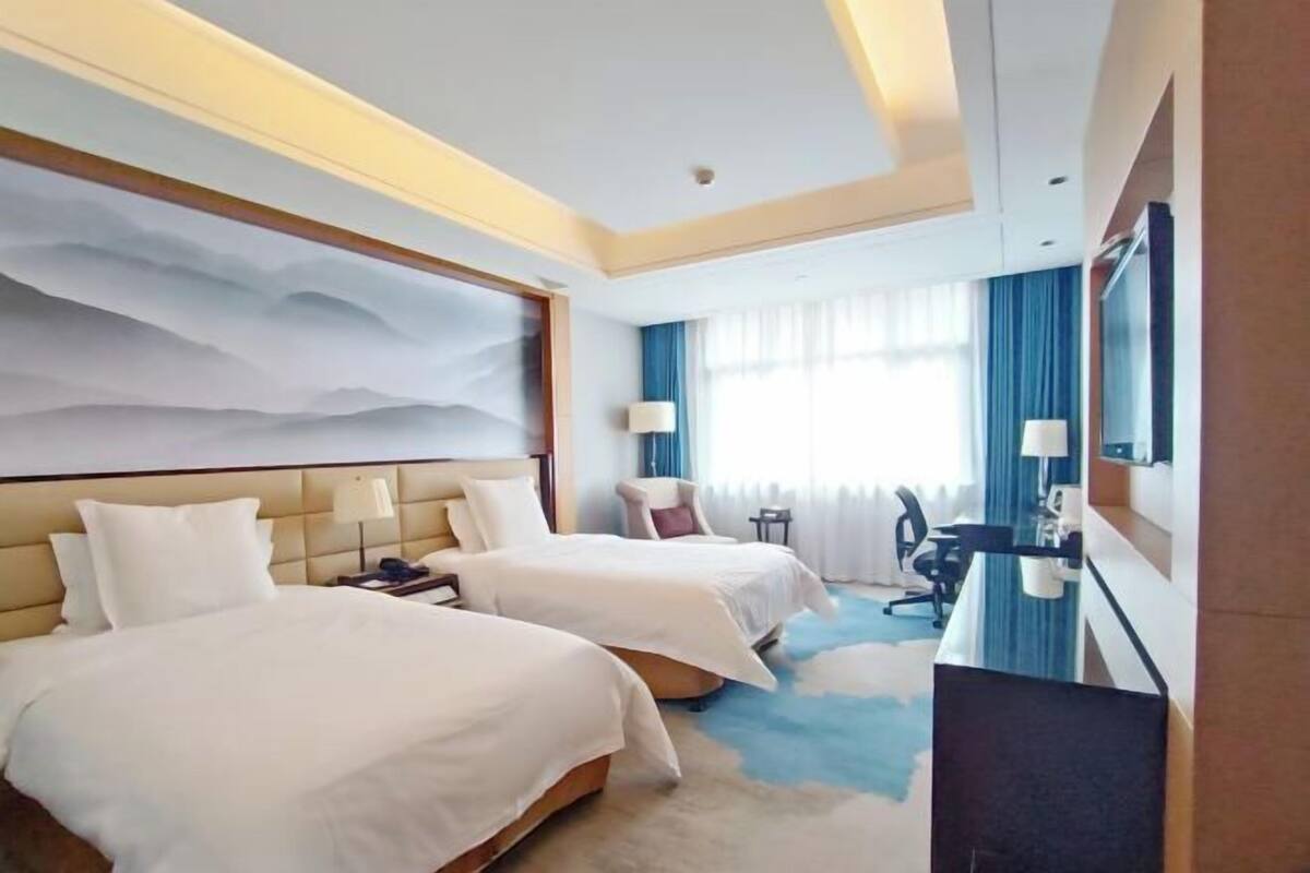 A hotel room with two beds, a desk, a chair, a TV, and a large window with curtains.