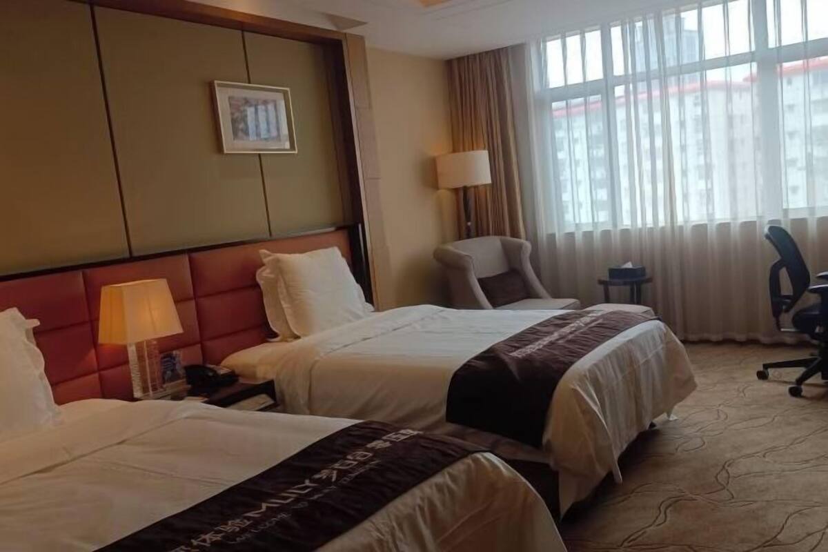 A hotel room with two beds, a headboard, a desk, a chair, a lamp, and a window with curtains.