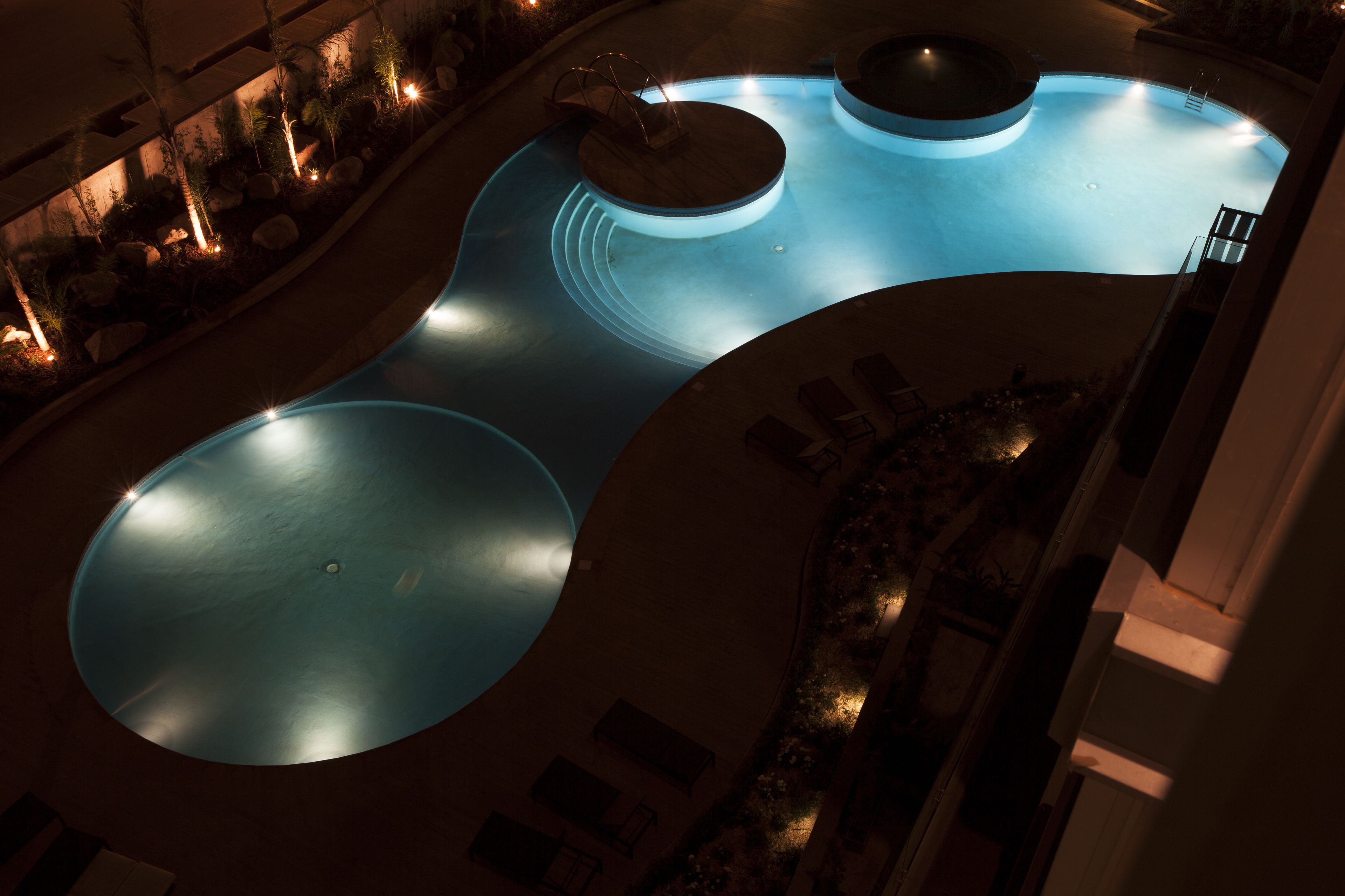 Una piscina de diseño circular, rodeada de sillas de descanso y un área ajardinada con rocas y plantas.