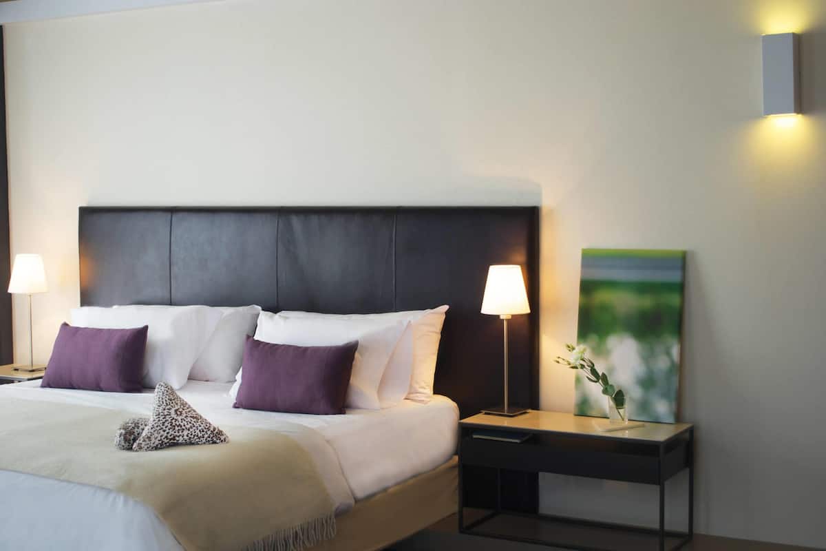 Cama bem arrumada com roupa de cama branca e roxa, cabeceira escura e duas luminárias de cabeceira.