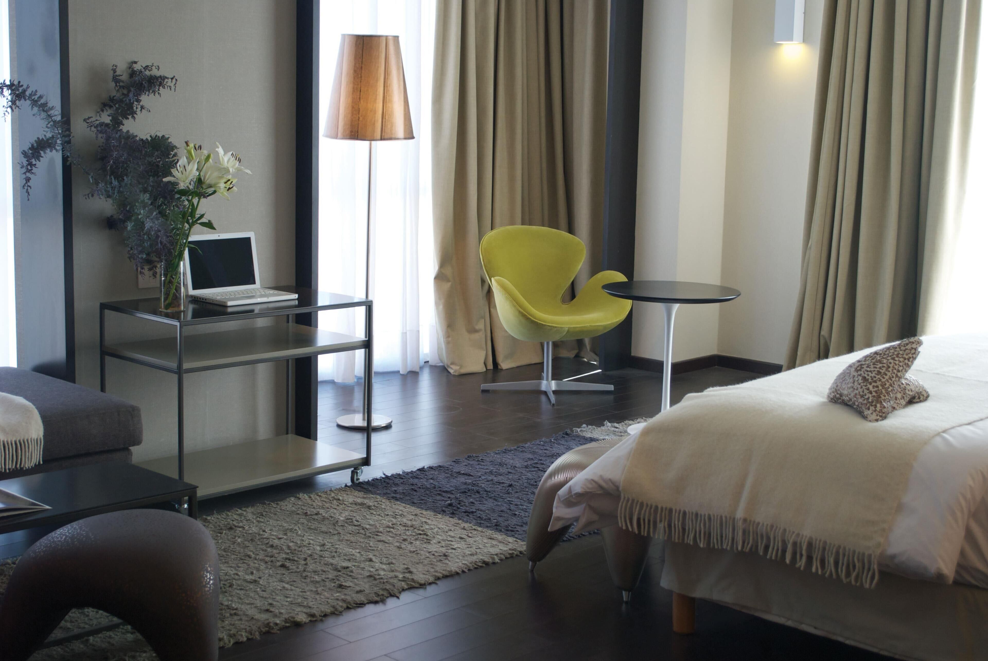 A modern hotel room with a bed, a desk with a laptop, a yellow chair, and a small table.