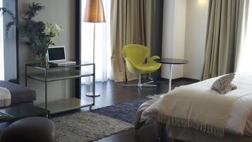 A modern hotel room with a bed, a desk with a laptop, a yellow chair, and a small table.