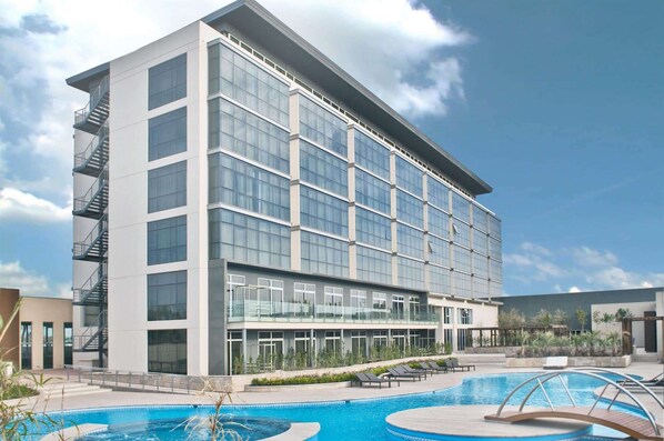 Een modern hotel met een groot zwembad, meerdere balkons en een heldere lucht.