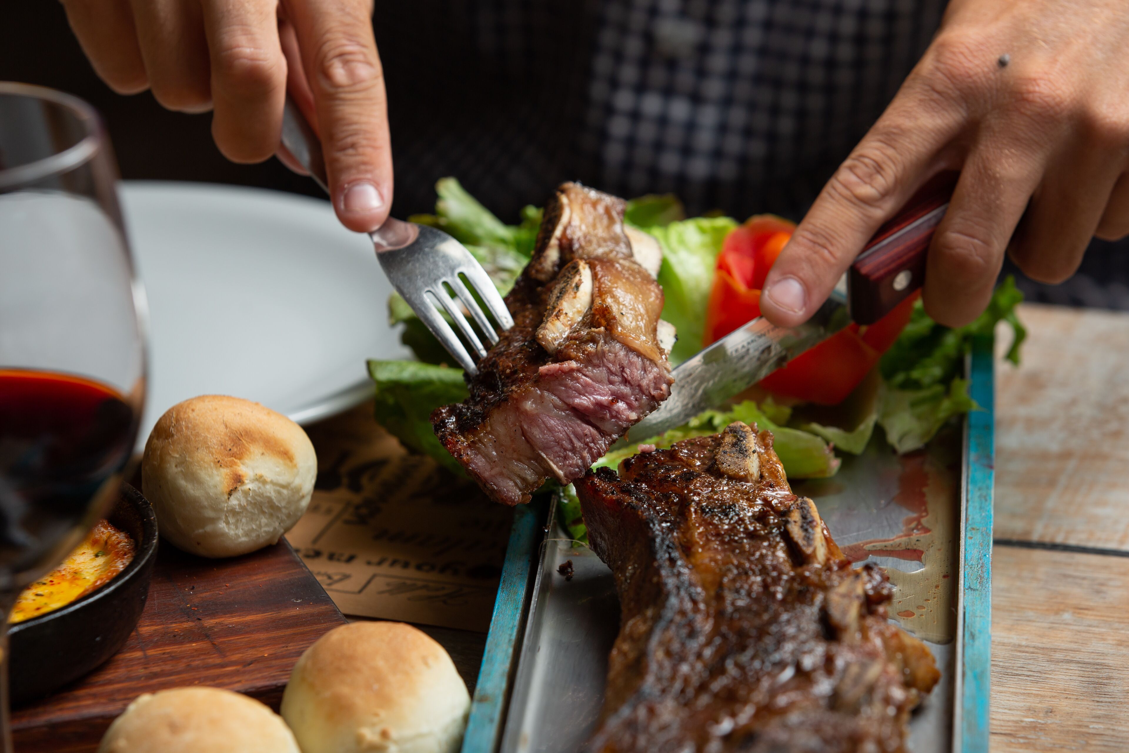 A person cutting a steak with a fork and knife on a plate with a side salad and bread rolls.