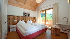 Ein Schlafzimmer mit Holzbalkendecke, einem Bett mit weißer Bettwäsche, einem Nachttisch, einem hölzernen Hocker, einem Waschbecken und einem großen Fenster mit Blick auf Grünflächen.