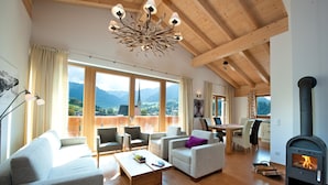 Ein geräumiges Wohnzimmer mit Holzboden, großen Fenstern mit Blick auf die Berge, einem Kamin und bequemen Sitzgelegenheiten.