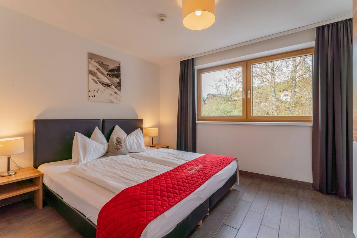 Ein Hotelzimmer mit einem großen Bett, einem Nachttisch mit Lampe, einem Fenster mit Vorhängen und einem Gemälde an der Wand.