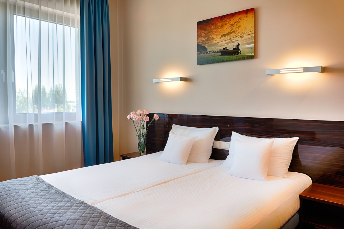 Una cama doble con sábanas blancas, un florero con flores rosas y un cuadro de atardecer en la pared.