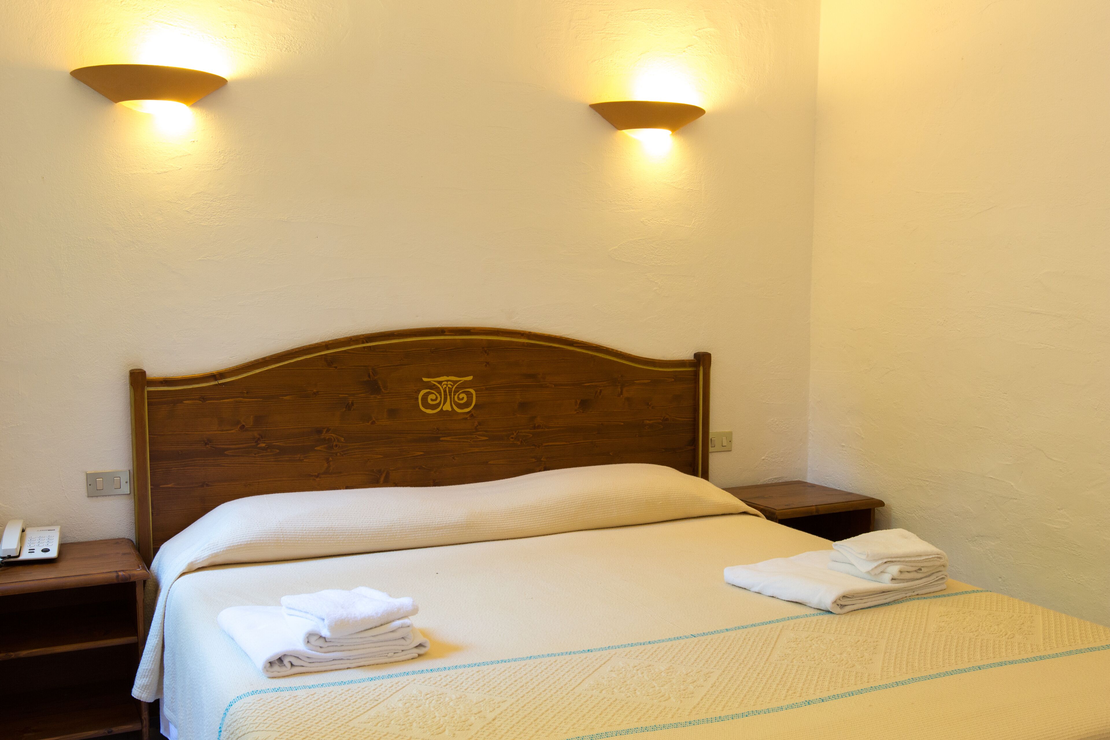 Una cama bien hecha con cabecera de madera, dos lámparas de noche y una toalla doblada sobre la mesita.