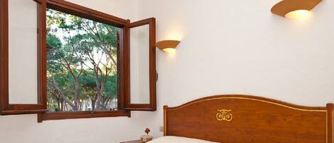 A bedroom with a wooden bed, a nightstand, a window with a view of trees, and wall-mounted lighting.
