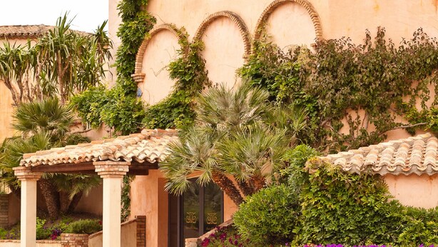 Un edificio con un patio cubierto, una pared de ladrillo y diversas plantas, entre ellas palmeras y arbustos florecientes.