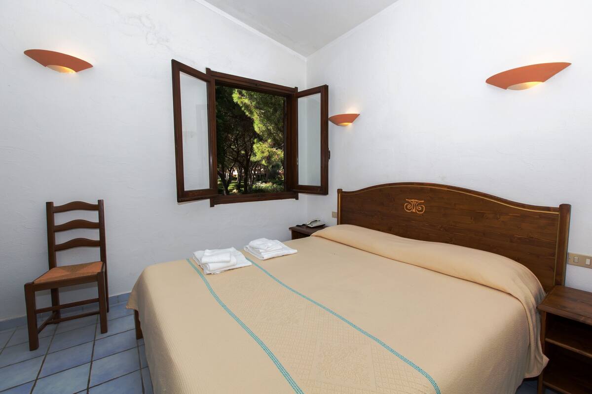 A neatly made bed with a beige bedspread and a blue accent, a wooden chair, a nightstand with a phone, and a window with a view of trees.