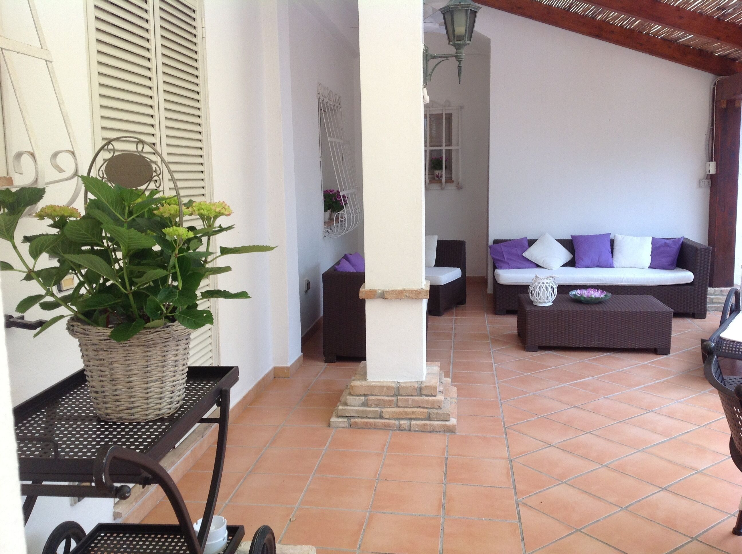 Un patio couvert comprenant une table en osier, un panier en osier avec une plante, un canapé en osier avec des coussins violets et une table basse en osier.