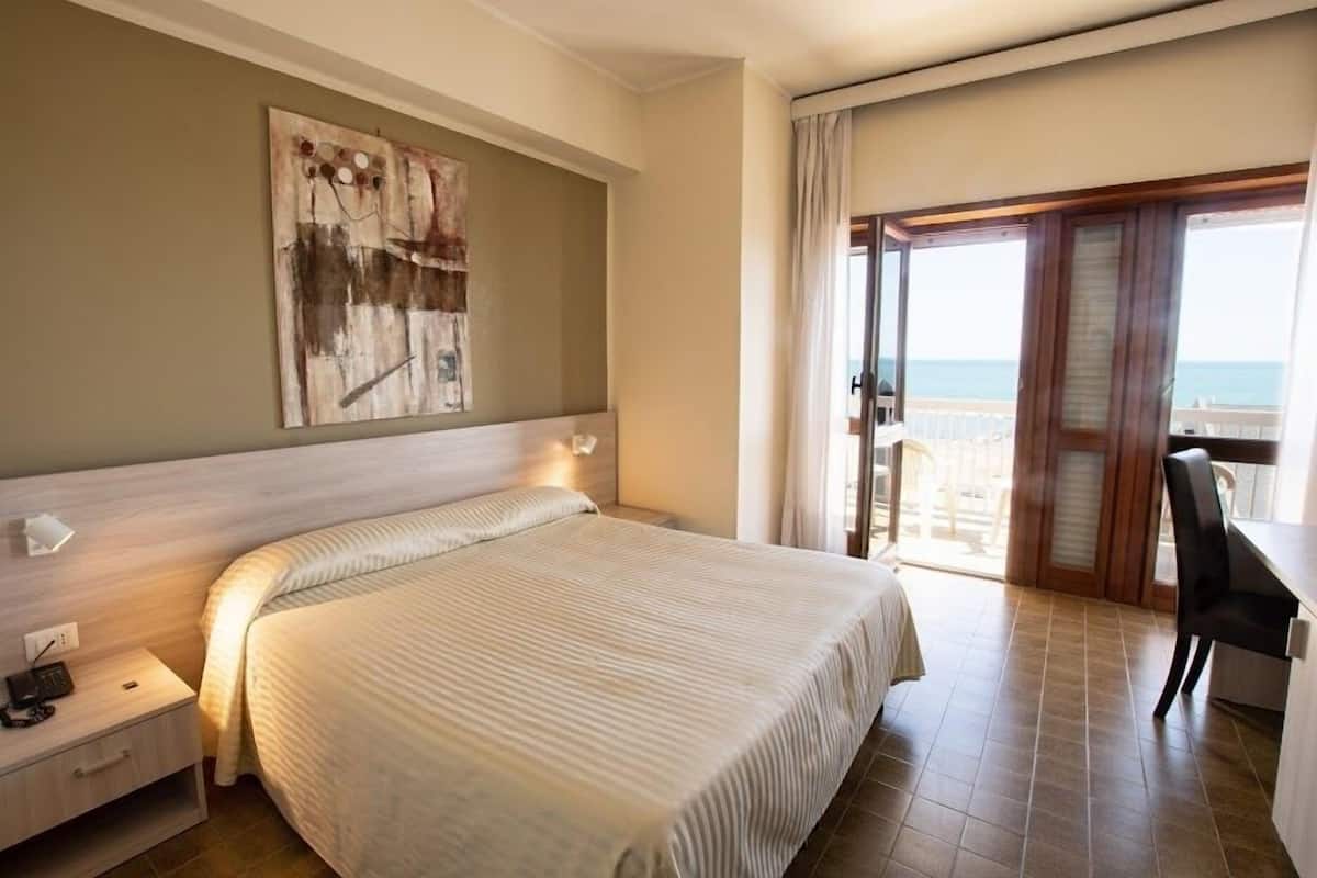 A hotel room with a large bed, a desk, a chair, a nightstand, and a view of the sea through a balcony.