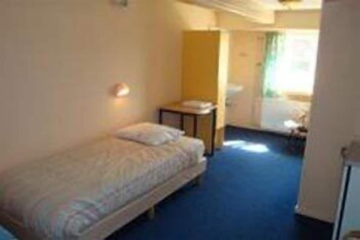 A small, single-bed room with a blue carpet, a bedside table, and a window.