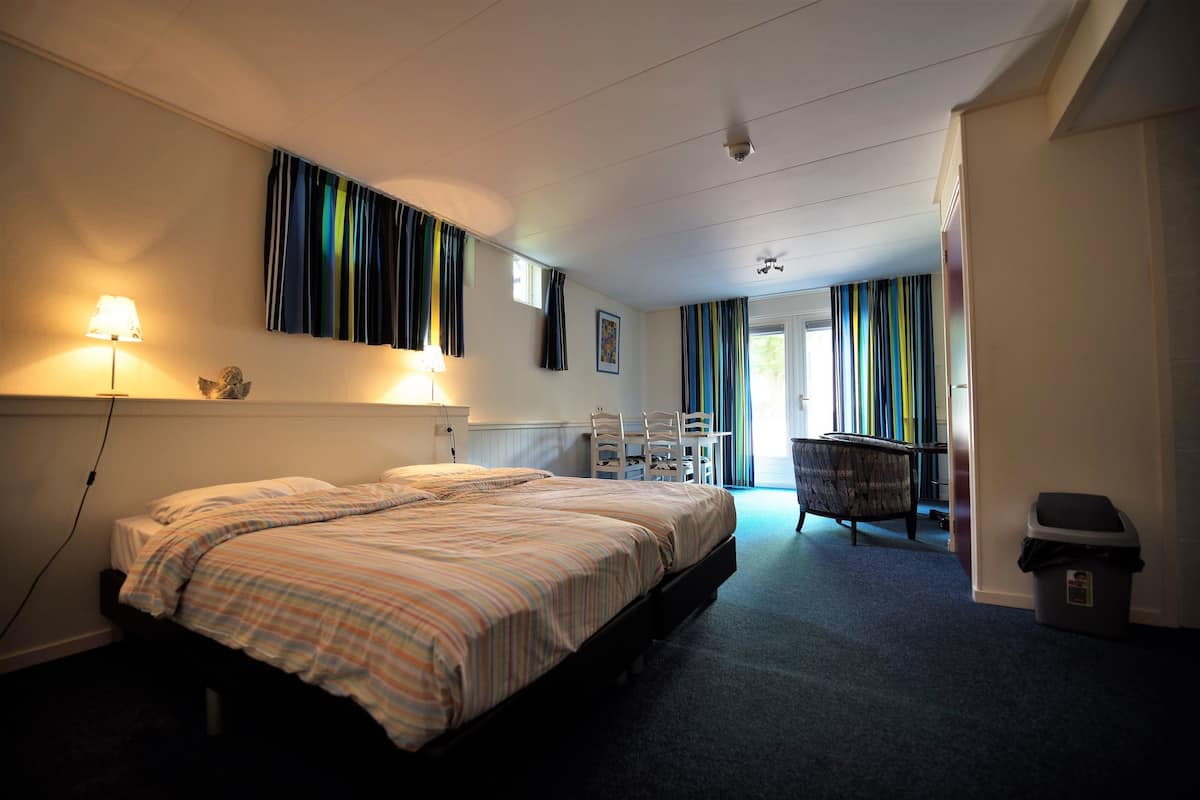 A hotel room with a large bed, a desk, and a chair. There is a window with curtains, a lamp, and a trash bin.