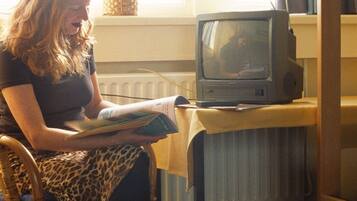 Una persona sentada en una silla leyendo un libro, con un televisor sobre una mesa frente a ella.