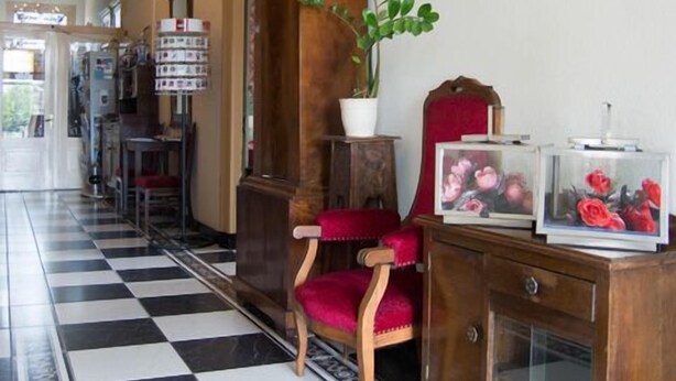 Un pasillo con suelo de cuadros, una silla de madera, un aparador con puertas de cristal y una lámpara fijada a la pared.