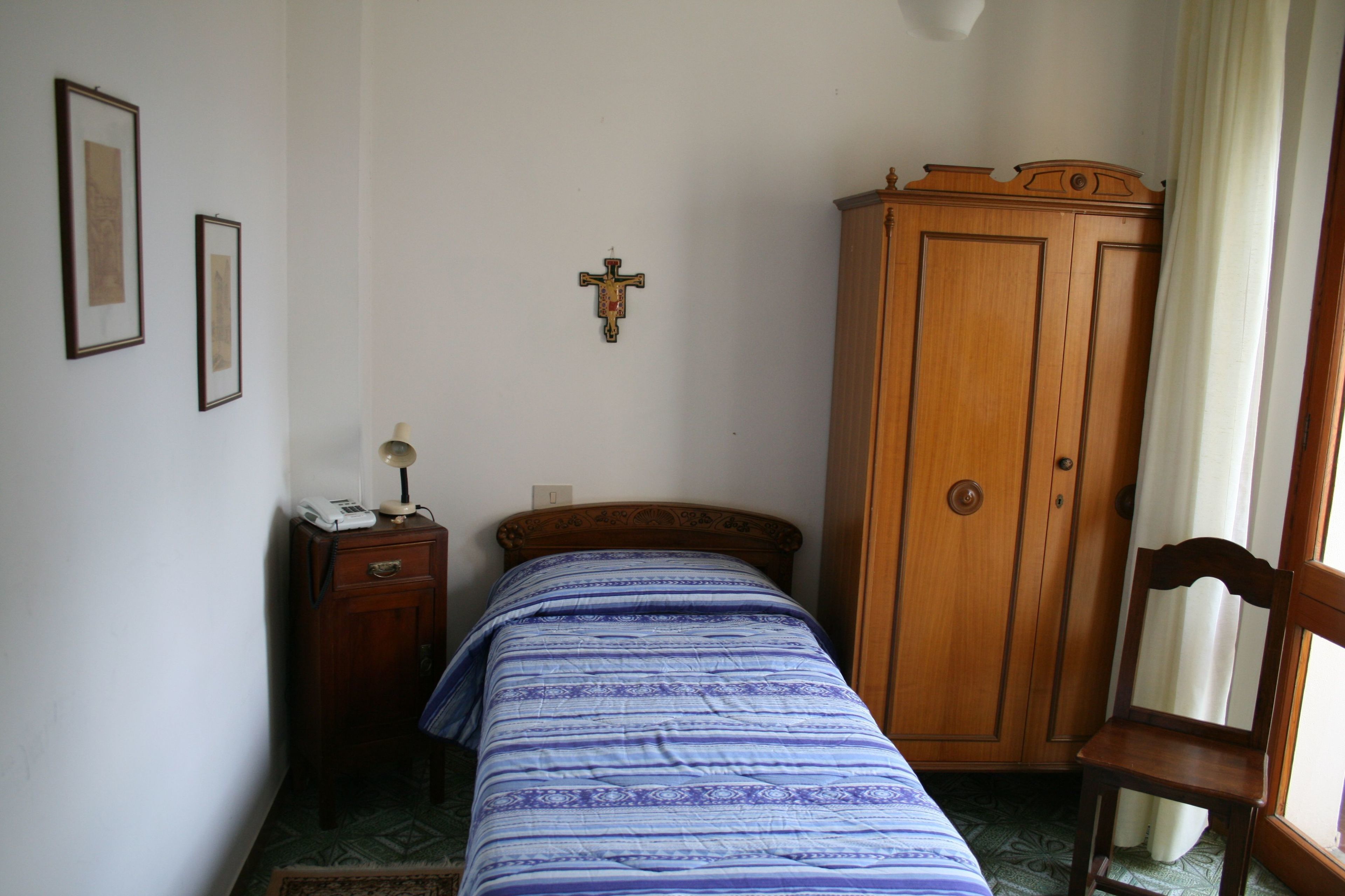 A bedroom with a single bed, a wooden wardrobe, a small bedside table with a lamp, and a framed picture on the wall.