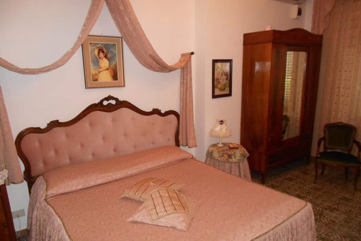 A bedroom with a large bed, a wooden wardrobe, a chair, and a small table with a lamp.