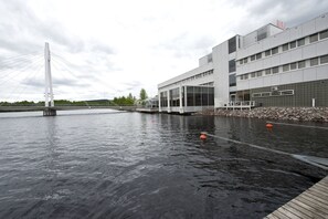 A modern building with a large glass facade, a bridge in the background, and a calm water body in the foreground.