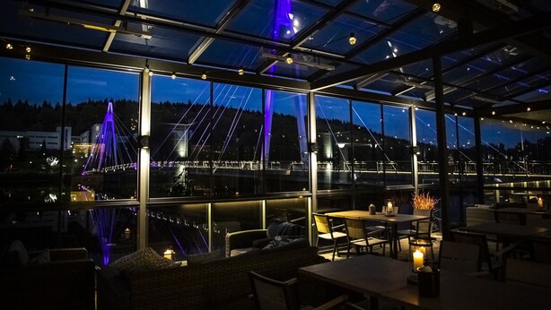 Un comedor interior con mesas y sillas, ventanas amplias y vistas a un puente con iluminación morada.