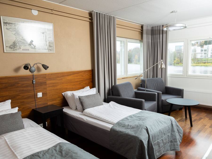 Ett hotellrum med en säng, två fåtöljer, ett litet bord, en lampa och ett fönster med utsikt över byggnader och en vattenyta.