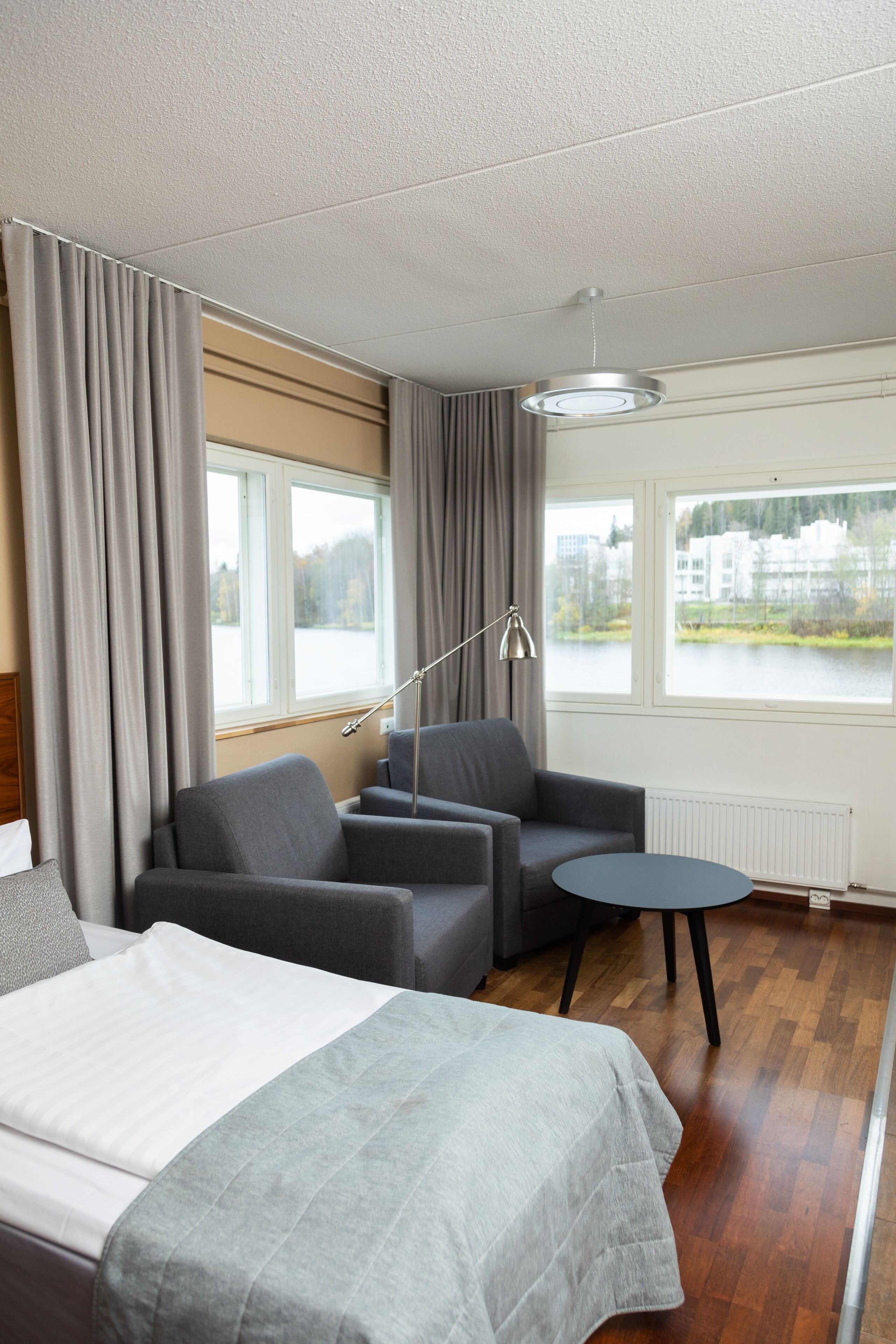 A modern hotel room with a bed, two armchairs, a small table, and a view of a lake and buildings through large windows.