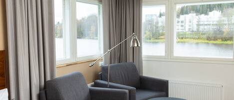 A modern hotel room with a bed, two armchairs, a small table, and a view of a lake and buildings through large windows.