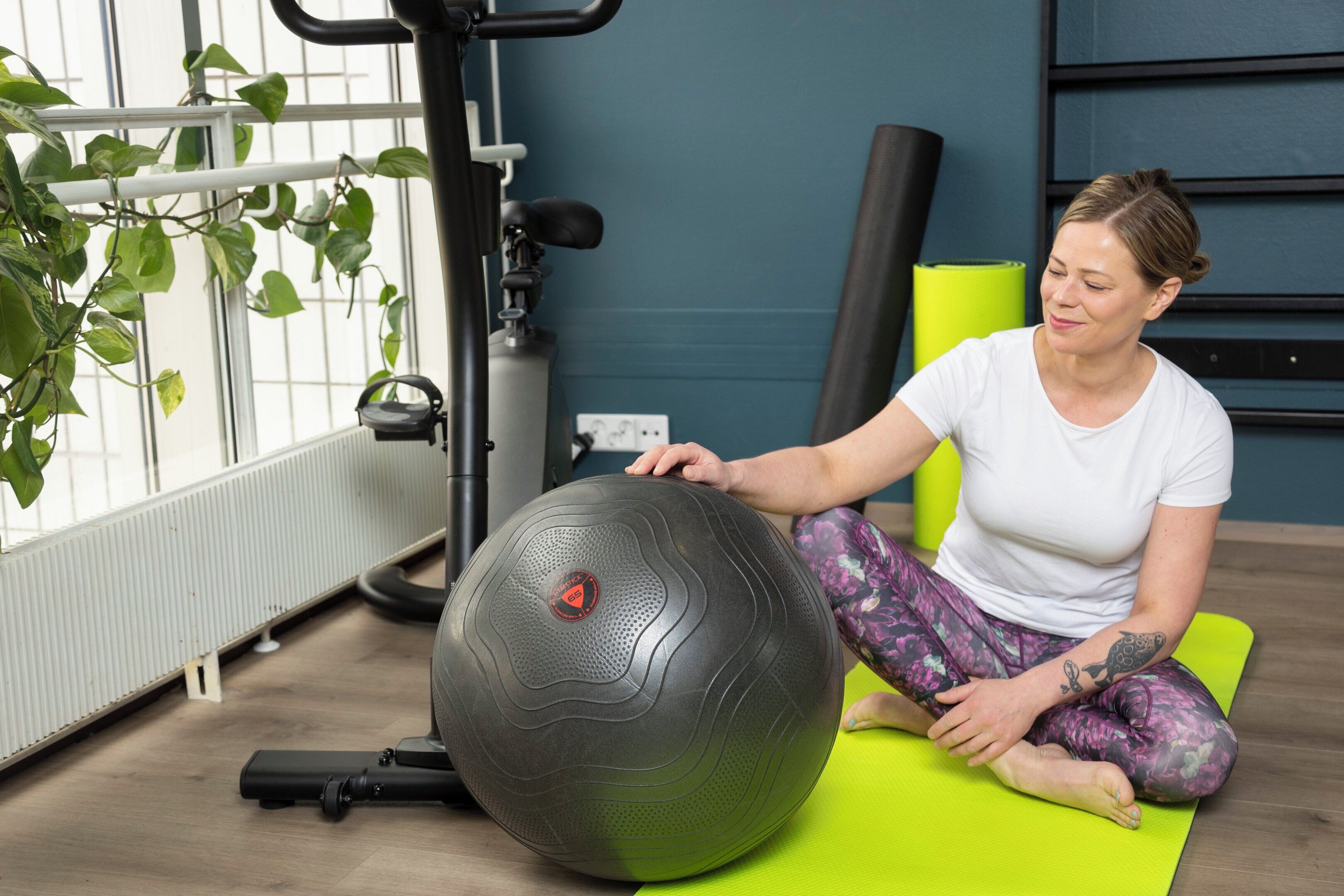 A person sitting on a green exercise mat in a room with exercise equipment, a radiator, and a window with plants.
