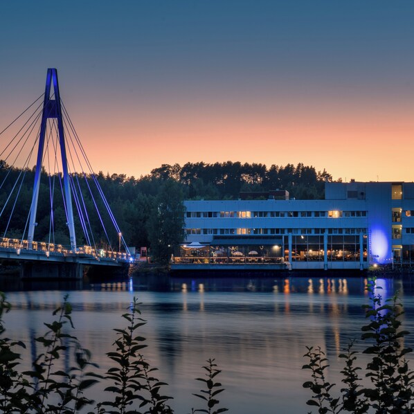 A modern building with a flat roof and large windows, situated by a calm body of water with a bridge in the background.
