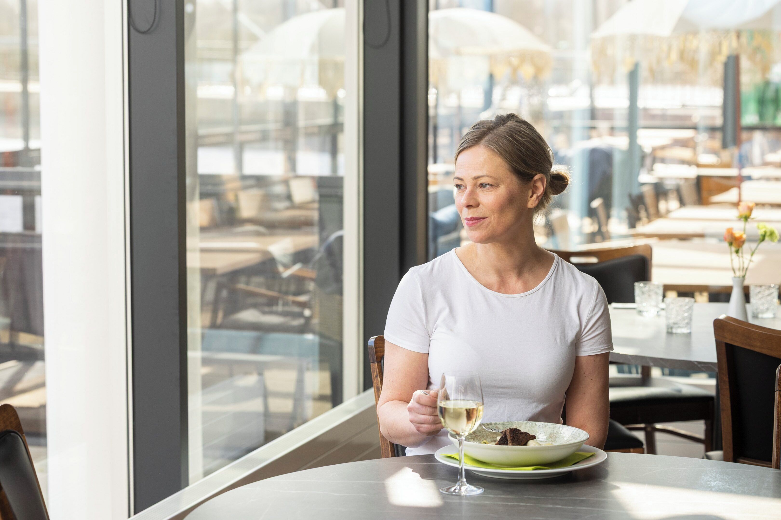 A person seated at a table with a plate of food and a glass of white wine, in a well-lit indoor space with large windows.