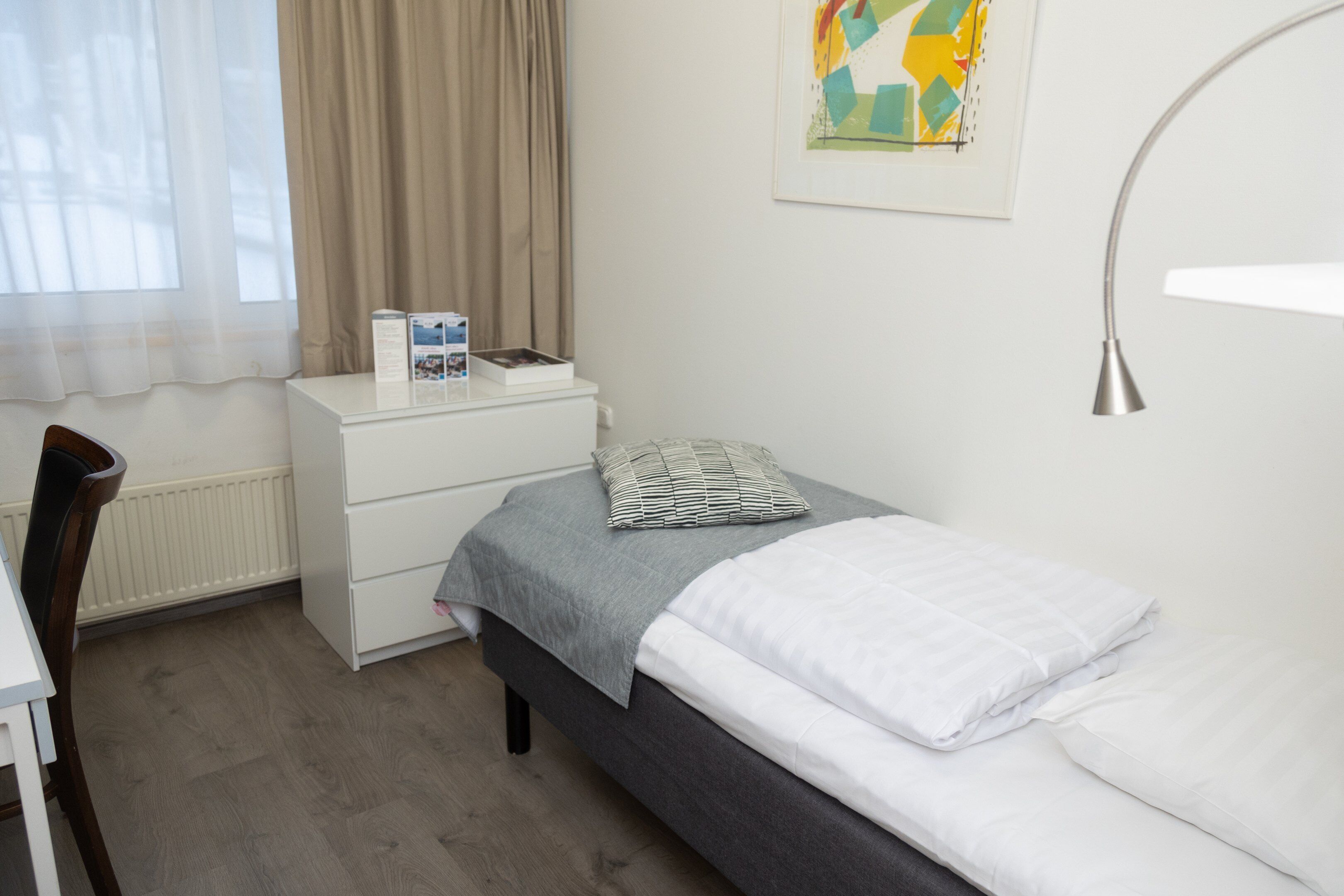Een slaapkamer met een bed, nachtkastje, stoel, bureau en een raam met gordijnen.