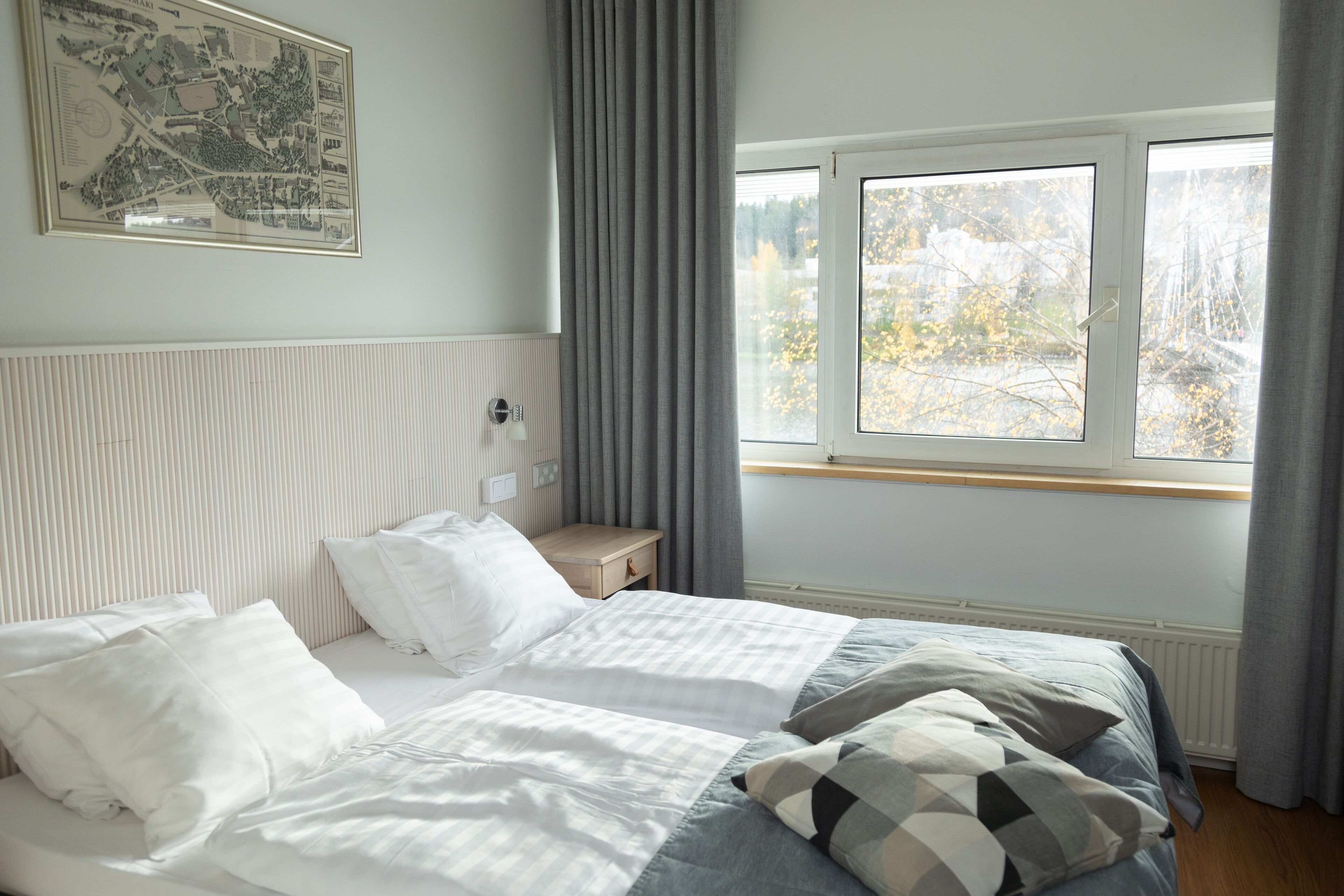A bedroom with two beds, a nightstand, a framed map on the wall, and a window with a view of trees.