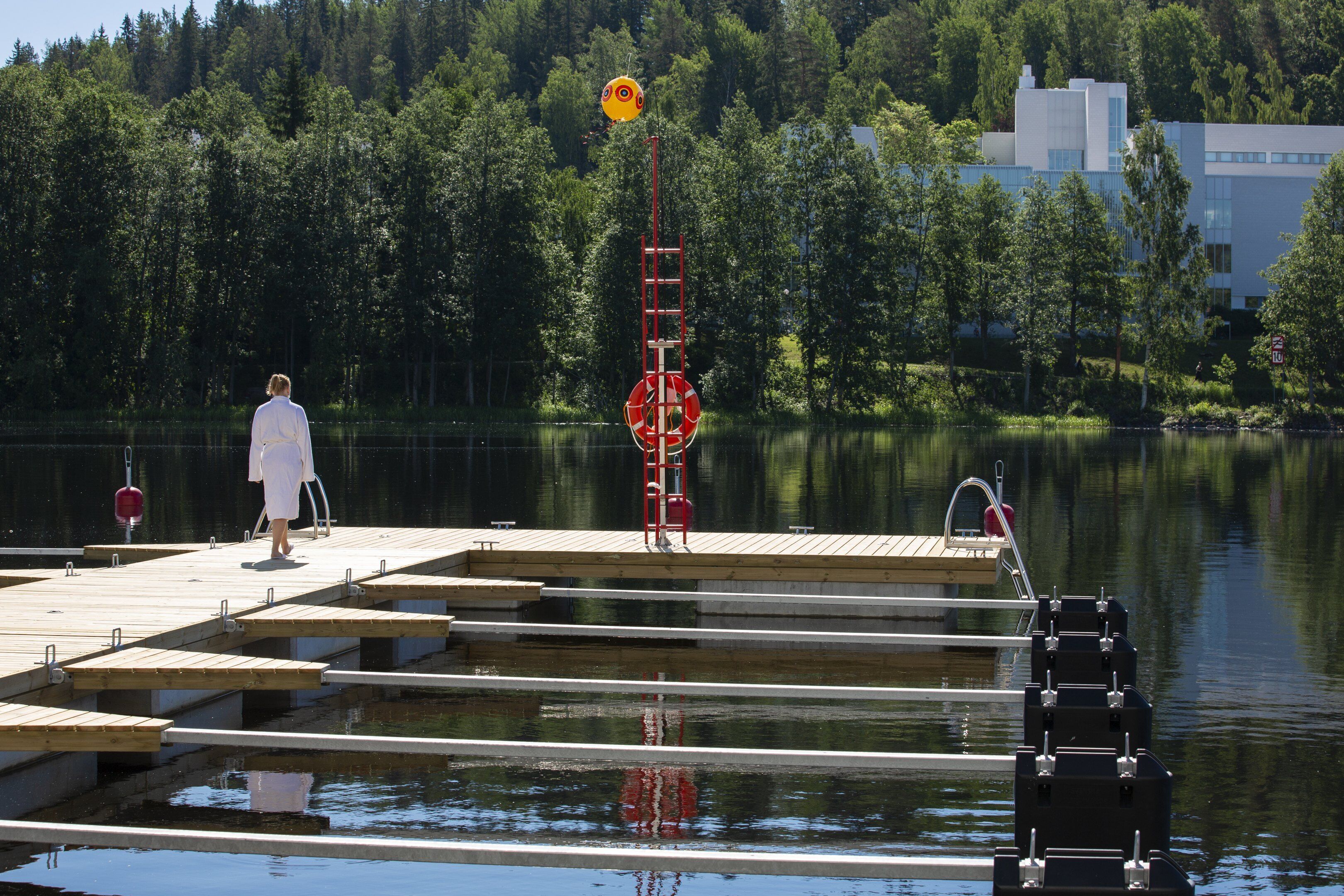 A wooden dock extending into a calm lake, surrounded by dense forest and a modern building in the background.