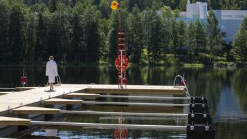 A wooden dock extending into a calm lake, surrounded by dense forest and a modern building in the background.