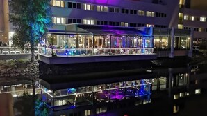 Un hotel moderno con una piscina reflectante en primer plano, que cuenta con un restaurante y mesas al aire libre, y un letrero visible que dice 'ALBA'.