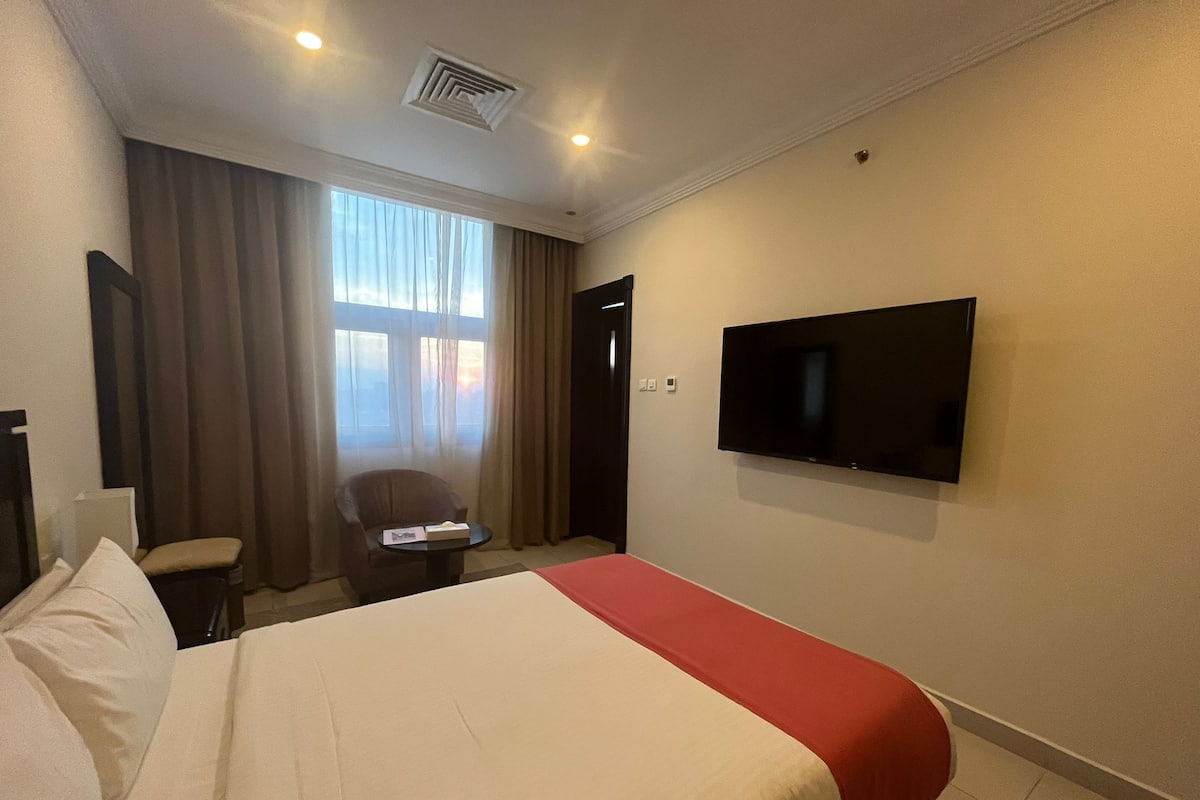 Une chambre d’hôtel avec un lit, une chaise, une table, un téléviseur et une fenêtre avec des rideaux.