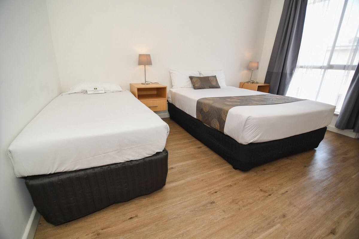 Ein Hotelzimmer mit zwei Einzelbetten, Holzboden, Nachttischen mit Lampen und einem großen Fenster mit Vorhängen.